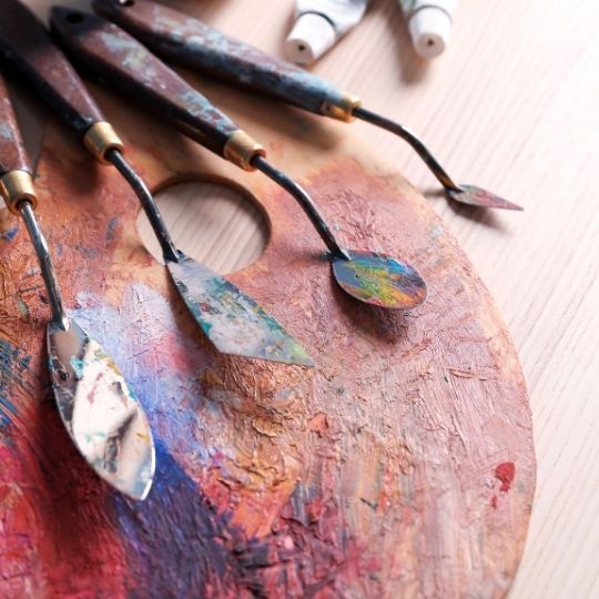 Espátulas artísticas sobre paleta de pintura colorida, inspirando texturas para Sphata, a cerâmica com efeito gesso e cimento da Recer